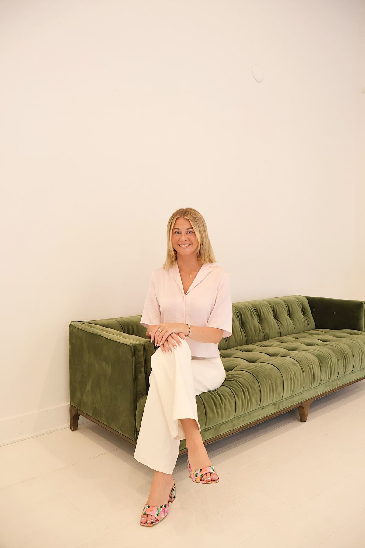 Nat smiling, seated on a green velvet sofa in a blush top, white pants and floral heels