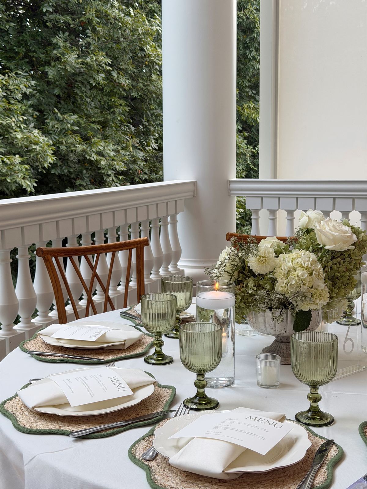 Round table set with green glassware, woven placemats, menus, and a white and green floral centerpiece