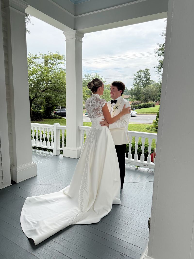 Bride in a lace-back gown beside a groom in a white dinner jacket on a porch