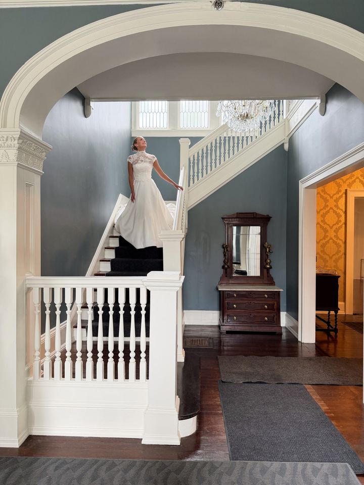 Bride in a high-neck lace gown on a white staircase, looking up at a chandelier