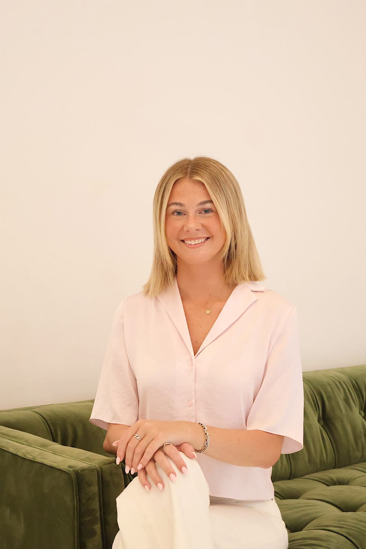 Nat smiling, seated on a green velvet sofa in a blush top